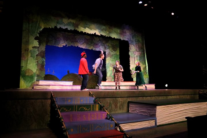 Four actors perform on a colorful stage with a backdrop resembling a whimsical forest. The stage includes large, book-like steps leading up to the platform, with a painted blue sky and wind turbines visible in the background. The performers appear engaged in an animated scene under dramatic lighting.